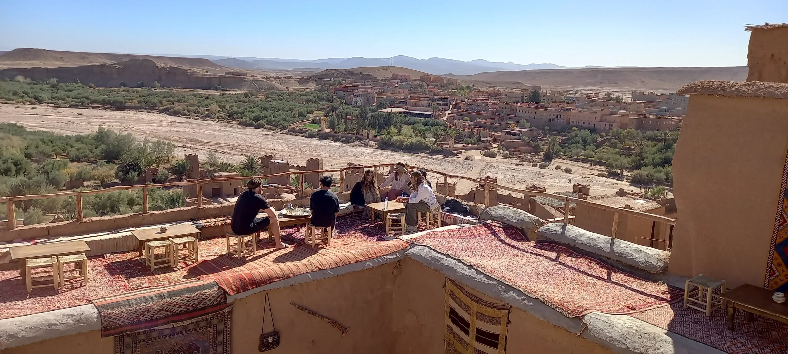 English-speaking travelers enjoying mint tea with a local Berber guide on a rooftop terrace overlooking a desert village in southern Morocco.